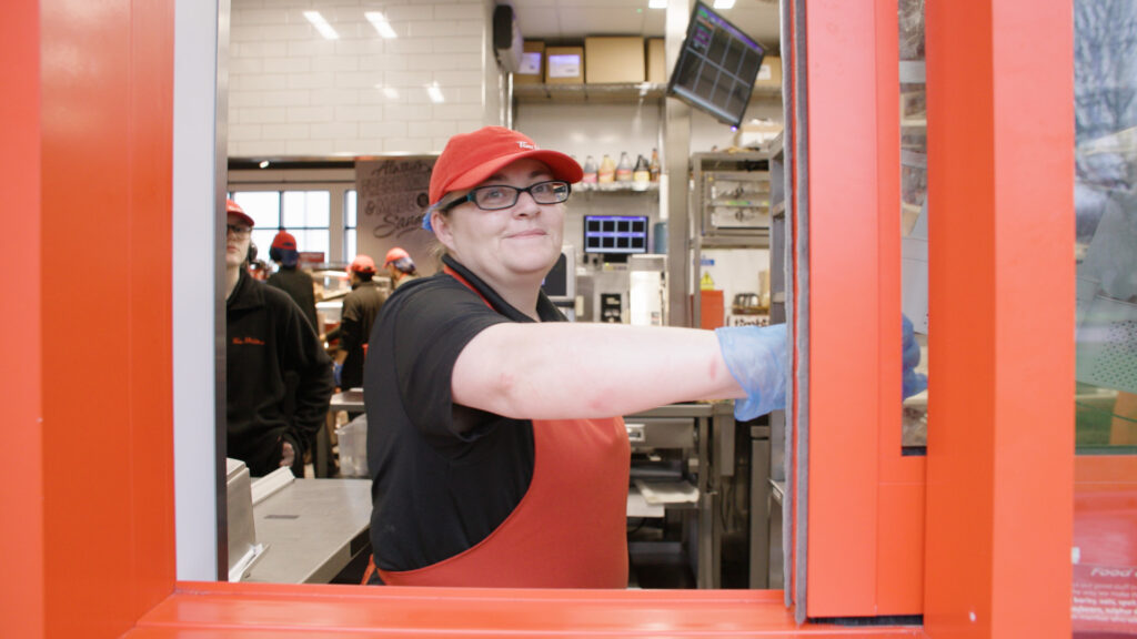 Tim Hortons team member stands at drive through window. She is wearing a red apron and smiling, closing the window.